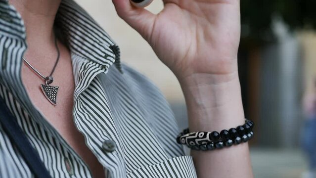 Close-up of a woman with a triangular silver pendant on her chest and a bracelet made of stones on her wrist is talking on the phone. Stylish woman in the city