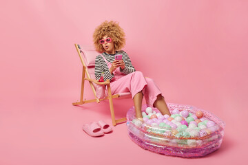 Surprised woman uses mobile phone for chatting online looks wondered aside enjoys summer vacations keeps legs in inflated pool poses on deck chair isolated over pink background. Holidays concept