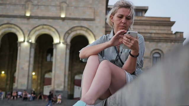 A young adult Caucasian woman against the background of a beautiful theater building in a long striped dress and beautiful legs sits on the parapet and uses a smartphone with her fingers