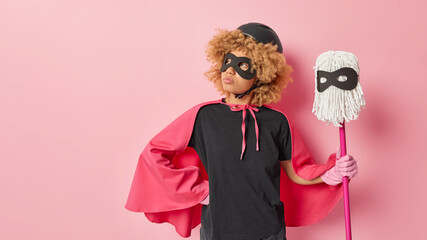 Horizontal shot of thoughtful woman keeps hand on waist dressed in superhero costume holds mop for...