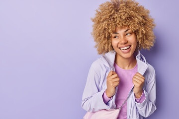 Horizontal shot of cheerful young European woman with curly hair wears jacket carries bag smiles happily focused away isolated over purple background copy space for your advertising content.