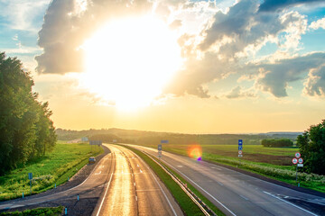 summer landscape, highway with road signs shining with reflected light with glare from the bright, blinding sunset orange sun shining through a lot of clouds