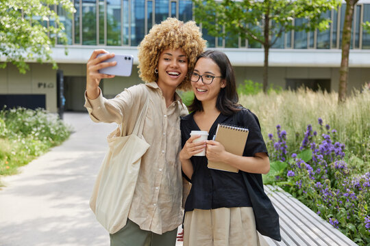 Outdoor shot of two female students stroll at campus take selfie via smartphone carries spiral notebook and takeaway coffee dressed casually make memorable photo together. Friendship and lifestyle