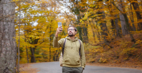Obraz premium Young handsome man in autumn forest with smartphone make photo. hipster guy with backpack , traveller standing in woods, Hiking, Forest, Journey, active healthy lifestyle, adventure, vacation concept.