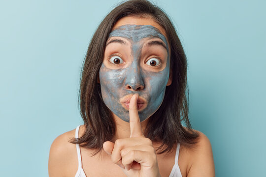 Mysterious Asian Woman With Dark Hair Applies Clay Facial Mask To Reduce Signs Of Agening Like Fine Lines Wrinkles Or Brown Spots Makes Hush Gesture Tells Secret Of Beauty Isolated Over Blue Wall