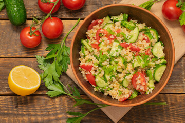 Tabbouleh. Vegetarian salad with bulgur, tomatoes, cucumbers, parsley, lemon juice, olive oil. Tabbouleh on a wooden table with of fresh vegetables, view from above, top view.