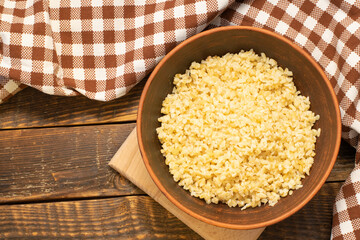 Cooked bulgur porridge in a clay bowl on a wooden table with checkered kitchen towel. Bulgur, a cereal made from wheat grains.. Top view, space for text, copy space.