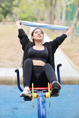 Obraz premium Happy smiling young beautiful asian woman in sportswear relaxing on public exercise machine. Cute and sporty girl working out on bike and using mobile smartphone outdoor