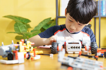 Smart looking Asian student kid assembling robot, coding and solving engineering problem at home with yellow wall in background. STEM education and 21st century learning skills concept
