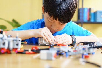 Smart looking Asian student kid assembling robot, coding and solving engineering problem at home with yellow wall in background. STEM education and 21st century learning skills concept