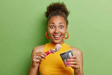 Positive surprised woman with hair bun eats cold fresh ice cream with spoon looks gladfully at camera wears earrings and casual yellow t shirt has upbeat mood isolated over green background. - Powered by Adobe