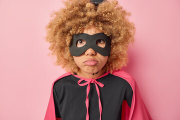 Unhappy curly haired woman wears black eyemask t shirt and cloak feels very sad pretends being hero and having supernatural power isolated over pink background. Fictional character fights for good