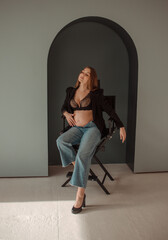 pregnancy fashion. young pregnant girl in black jacket and jeans is sitting in director's chair on a mint wall background with arch and looking apart with hand near belly. pregnant concept, free space