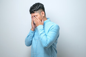 Asian young man with mustache wearing casual shirt with sad expression covering face with hands while crying.