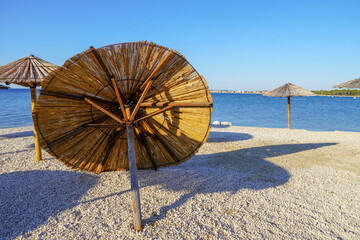 A hat, a straw beach umbrella, removed and resting on its stand, stands in a row on a sandy beach with azure sea water. End of the beach season
