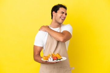 Restaurant waiter holding waffles over isolated yellow background suffering from pain in shoulder...