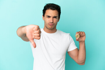 Young man holding a Bitcoin over isolated blue background showing thumb down with negative...