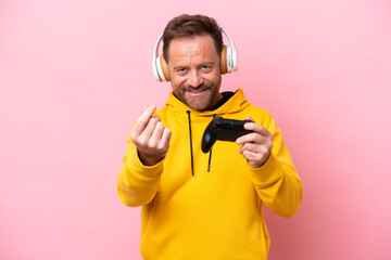 Middle age man playing with a video game controller isolated on pink background making money gesture