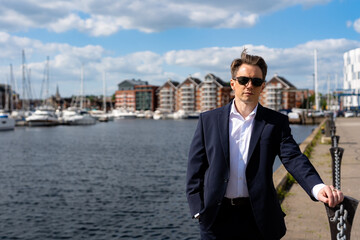 A handsome businessman in a casual dark blue suit in front of a busy marina with luxury yachts moored up