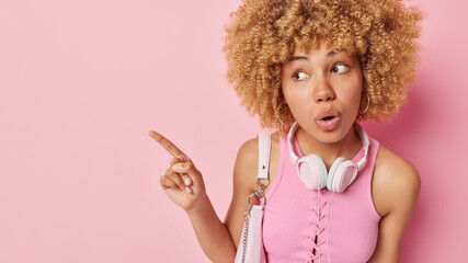Stunned curly haired young woman dressed casually poses with bag and stereo headphones around neck points away at blank space has shocked expression isolated over pink background. Omg look there