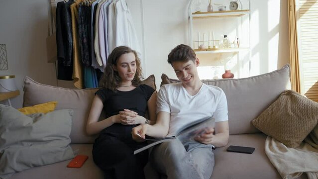 Woman tells funny stories watching school album with partner on weekend. Young couple sits on couch in living room at home remembering moments