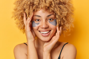 Headshot of positive young woman with curly hair applies beauty patches under eyes to reduce puffiness smiles toothily has healthy smooth skin isolated over yellow background. Wellness concept