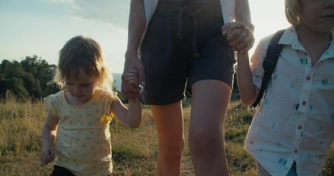 Family together hike on summertime travel adventure holding hands. Mother with children walk on beautiful nature landscape at evening. Sister and brother hold mum hand.