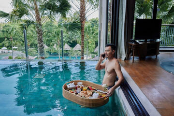 Happy man in swimsuit having floating breakfast tray in luxury pool hotel while talking on the...