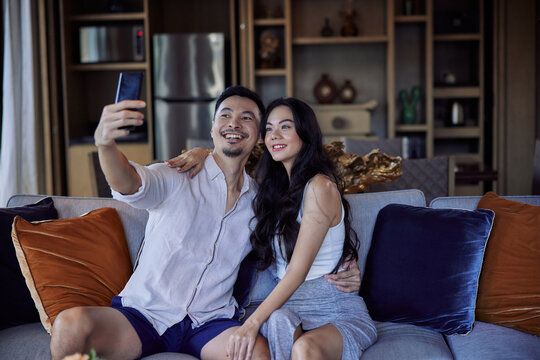 Husband and wife taking selfie photo in hotel room after check-in. Happy asian couple with suitcases having fun in living room of luxury villa