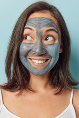 Positive dark haired young woman with eastern appearance applies beauty mask to reduce skin imperfections and provide relaxation focused on left has cheerful smile isolated over blue background