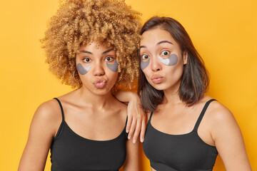 Obraz na płótnie Canvas Horizontal shot of surprised mixed race young women keep lips folded apply grey hydrogel patches under eyes to reduce wrinkles dressed in casual black t shirts isolated over yellow background