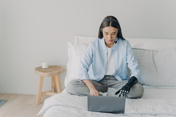 Disabled european woman with artificial arm is working on laptop in bedroom sitting on her bed.
