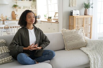 Biracial young girl enjoy meditation, practice yoga, sitting on sofa in lotus pose at home