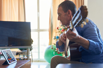Activity at old age concept. Senior male watching online guitar classical lesson with fitball and christmas tree on background