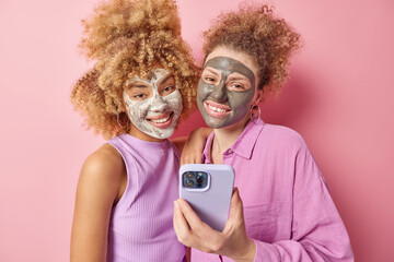 Positive young female friends with curly hair applies beauty mask for skin treatment use mobile phone dressed in casual clothes take selfie isolated over pink background. Facial care concept