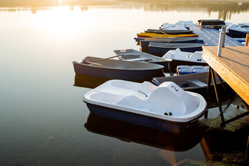 Fototapeta na wymiar Calm water boat dock, morning dawn glare of the sun, white catamaran for river walks, boat rental in the park, city pond.
