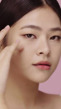 Vertical video close up face of beautiful young Asian woman with clear skin lifting eye with finger on pink background.
