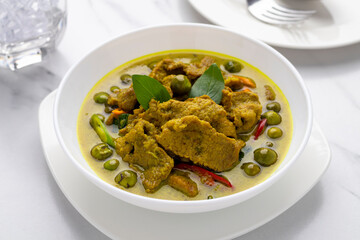 Thai green curry pork slices with pea eggplants served in a white serving bowl. This light yellowish green curry is one of popular Thailand’s signature dishes popularly served in Thai restaurants.