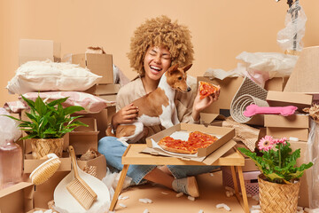 Positive curly haired woman poses with dog eats tasty pizza during relocation sits crossed legs dressed in casual clothes surrounded by cardboard boxes with household items. Moving day concept