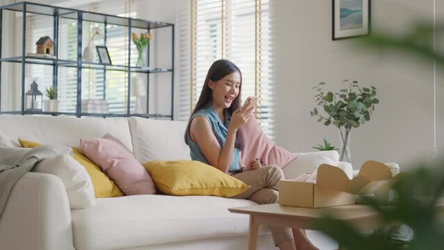 Gen Z micro influencer girl open mail box gift sit at home sofa couch. Asia people enjoy summer sale unbox buy order at online retail store shoot photo post show share on reel instagram social media.