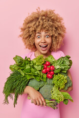 Surprised cheerful woman with curly hair carries fresh vegetables bought on market impressed to...