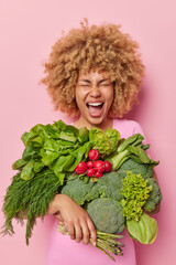 Indoor shot of curly haired young woman carries fresh green vegetables being vegetarian eats...