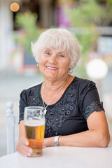 Senior woman sitting at a table in a summer cafe and drinking beer from a tall glass