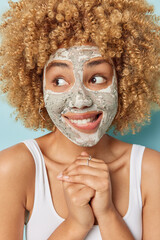 Pleased curly haired young woman applies nourishing clay mask bites lips keeps hands together looks away dressed in casual t shirt isolated over blue background. Skin care and wellness concept