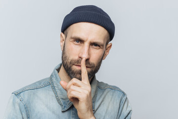 Headshot of handsome man with thick beard and mustache, has secret look, demonstrates silence sign,...