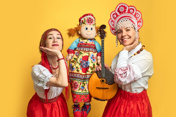 Women musicians in Russian folk dresses with musical instruments on a yellow studio background. Happy artists from Russia in national clothes with stringed musical instruments with smiles on faces