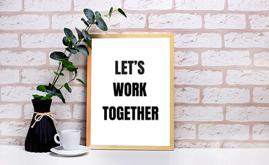 On a white table against a light brick wall, a branch of eucalyptus in a dark vase, a white cup and a light wooden frame with the text LET IS WORK TOGETHER. Home office interior.