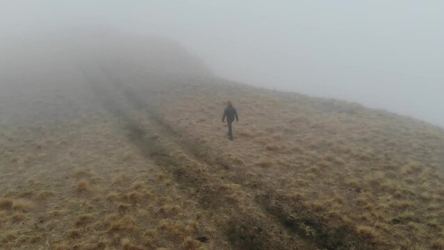 A caucasian man in tourist clothing walks on the top of a plateau next to a deep cliff along with a drone in fog and low cloud cover. aerial view