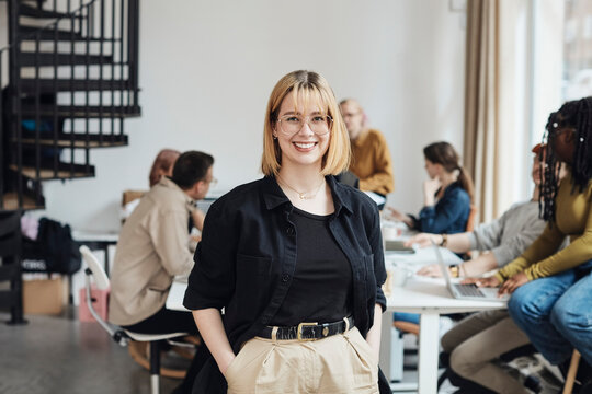 Portrait Of Smiling Young Female Computer Programmer Standing With Hands In Pockets At Tech Start-up Office