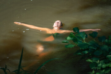 Fototapeta na wymiar A young beautiful dark-haired girl swims in a pond in the shade of trees.